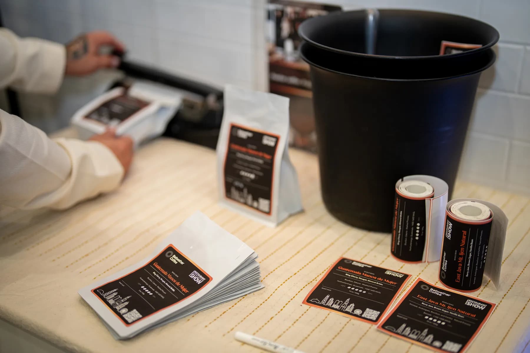 Printed labels, label rolls, and a labelled bag on a counter, with a bag being sealed behind them
